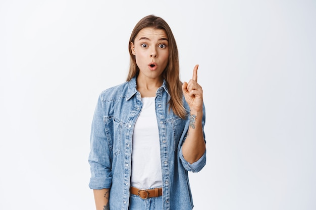 Chica rubia emocionada levantando el dedo en el signo de eureka y jadeando, sugiriendo un gran plan, encontró una solución, de pie contra la pared blanca