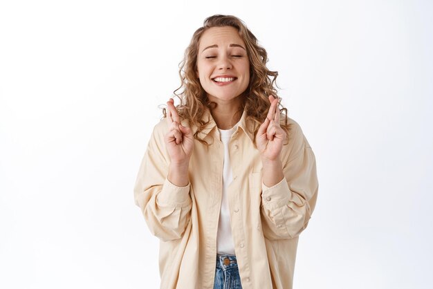 Una chica rubia emocionada que desea esperar algo anticipando los resultados positivos del examen cruza los dedos para tener buena suerte con los ojos cerrados de fondo blanco