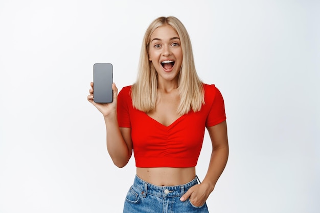 Chica rubia emocionada que muestra la interfaz de la aplicación de la pantalla del teléfono móvil o la aplicación de compras sonriendo asombrada de pie contra el fondo blanco