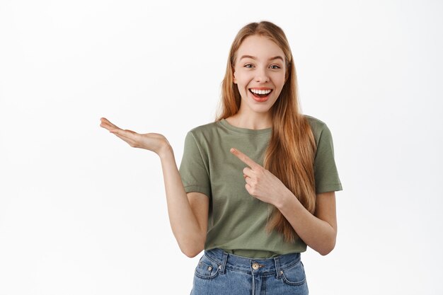 Chica rubia emocionada, sonriendo, apuntando a la mano abierta como si mostrara un artículo en la palma, recomendando un producto, exhibiendo publicidad, de pie contra la pared blanca