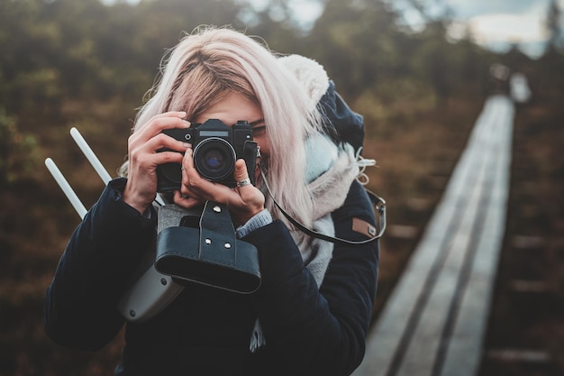 Chica rubia enfocada está haciendo una foto con su cámara de fotos mientras camina por el parque.