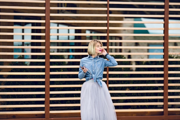 Chica rubia de ensueño con labios de color rosa brillante de pie y escuchando música en un teléfono inteligente con balks de madera a rayas detrás