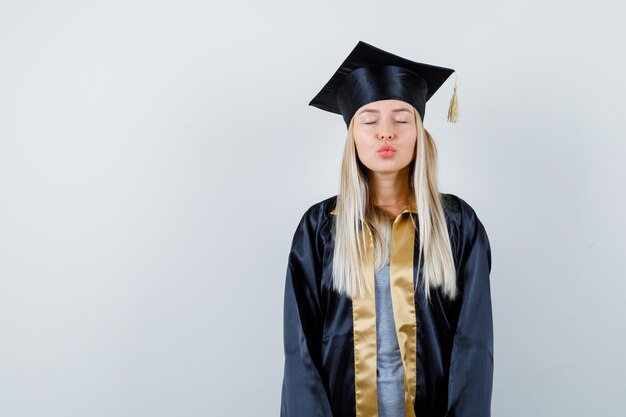 Chica rubia enviando besos en toga y gorra de graduación y luciendo linda.