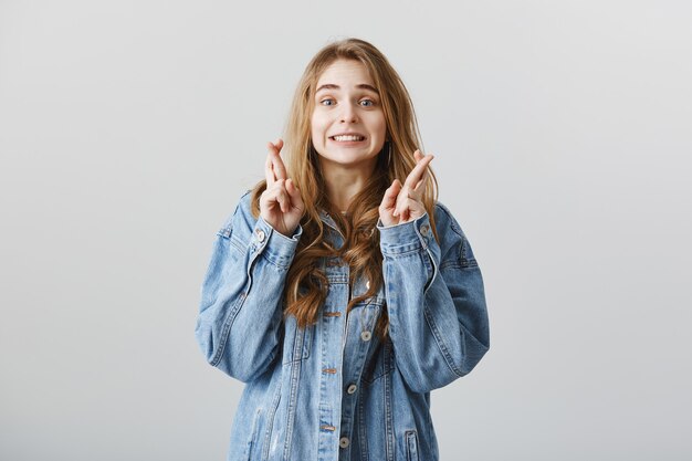 Chica rubia esperanzada en chaqueta de mezclilla, pidiendo deseo, cruzar los dedos como esperando resultados o noticias