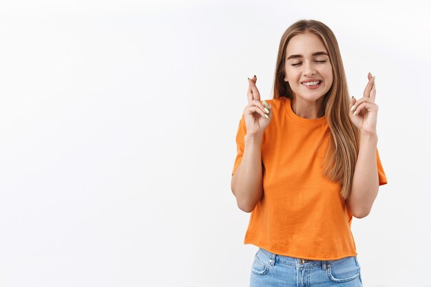 Chica rubia esperanzada pidiendo deseos, suplicando con todo su esfuerzo para que el sueño se haga realidad, cerrar los ojos y apretar los dientes con entusiasmo, pensar o soñar, cruzar los dedos, buena suerte, de pie en la pared blanca