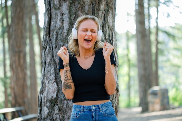 Chica rubia está gritando y levantando los puños mientras escucha música en los auriculares en el fondo de la naturaleza