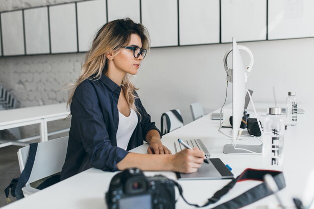 Chica rubia con estilo usando tableta después de la sesión de fotos y sentada en la mesa con computadora y cámara. Encantadora estudiante en gafas y camisa negra trabajando en proyecto.