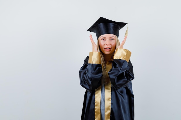 Chica rubia estirando las manos cerca de la cara en toga y gorra de graduación y mirando feliz.