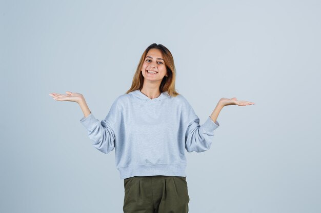 Chica rubia estirando las manos como sosteniendo y presentando algo en sudadera azul verde oliva y pantalones y mirando amable. vista frontal.