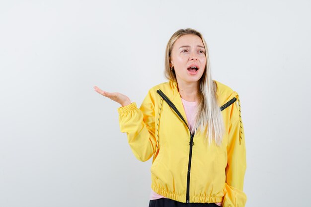 Chica rubia extendiendo la palma a un lado en chaqueta amarilla y mirando melancólica.