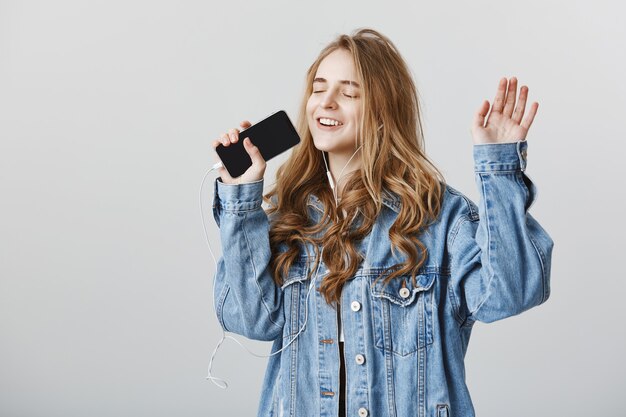 Chica rubia feliz despreocupada jugando aplicación de karaoke en el teléfono móvil, cantando en el teléfono inteligente con auriculares