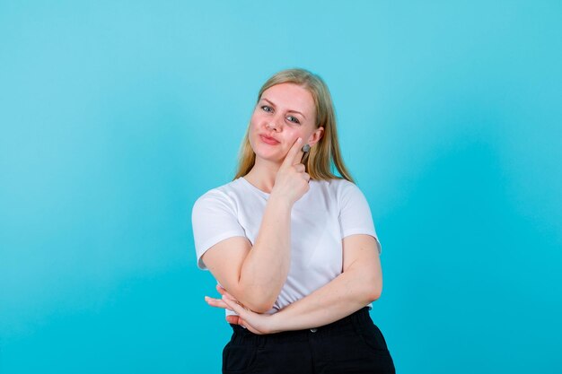 Chica rubia feliz está mirando a la cámara poniendo la mano en la mejilla sobre fondo azul.