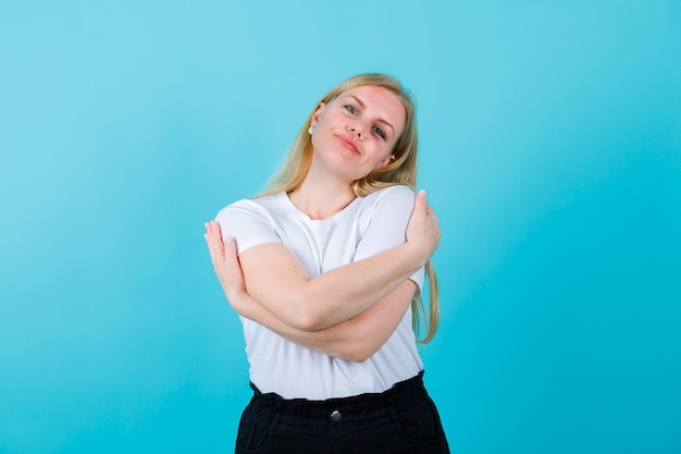 Chica rubia feliz está mirando hacia otro lado abrazándose a sí misma sobre fondo azul.