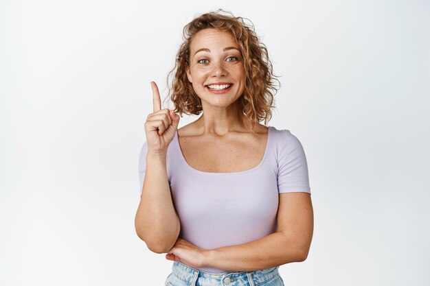 Chica rubia feliz señalando con el dedo hacia arriba, sonriendo y luciendo emocionada, mostrando publicidad con cara complacida, fondo blanco.