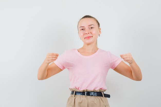 Chica rubia fingiendo sostener o mostrar algo en camiseta, pantalón y mirando alegre, vista frontal.