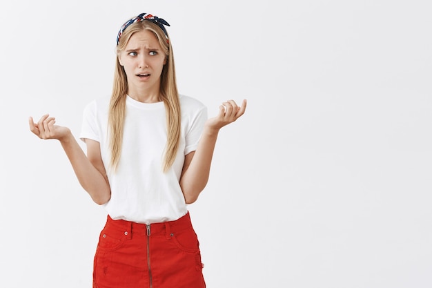 Chica rubia joven confundida y perpleja posando contra la pared blanca