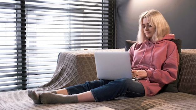 Chica rubia joven creadora de contenido está en su computadora portátil sentada en el sofá cerca de la ventana