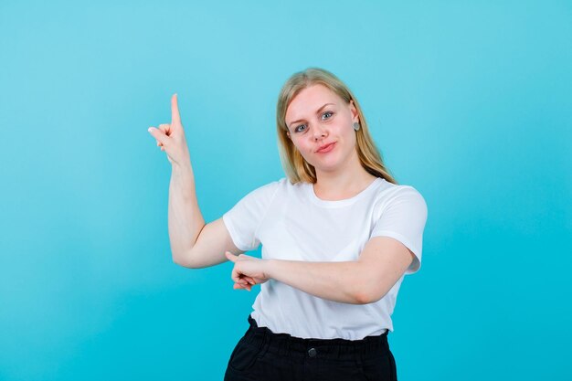 Chica rubia joven está apuntando hacia arriba con el dedo índice sobre fondo azul.