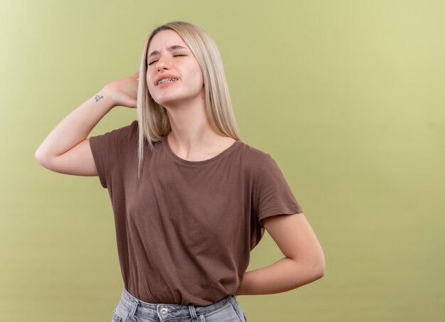 Chica rubia joven molesta en aparatos dentales poniendo las manos cerca de la cabeza y detrás de la espalda con los ojos cerrados en un espacio verde aislado con espacio de copia