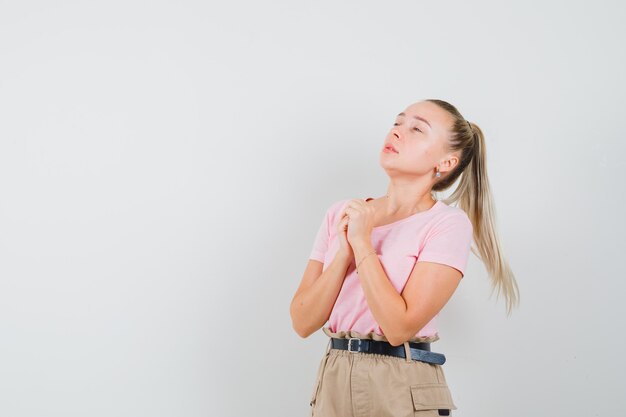 Chica rubia juntando las manos en gesto de oración en camiseta, pantalón y mirando esperanzado. vista frontal.