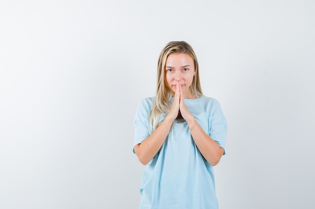 Chica rubia juntando las manos en posición de oración en camiseta azul y mirando bonita vista frontal.