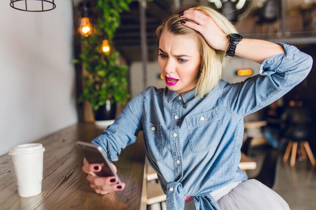 Chica rubia con labios rosados brillantes sentada en una cafetería tomando café y mirando su teléfono inteligente. Parece sorprendida y juega con su cabello. Viste camisa de mezclilla azul