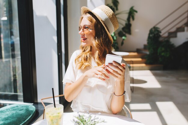 Foto gratuita chica rubia ligeramente bronceada mirando a otro lado con una sonrisa, sosteniendo el teléfono inteligente en las manos