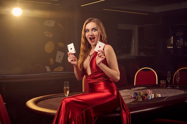 Una chica rubia llamativa con un vestido largo de satén rojo, con dos ases en las manos, se ve sorprendida y posa sentada en una mesa de póquer en un casino de lujo. Pasión, cartas, fichas, alcohol, ganar, apostar - es un
