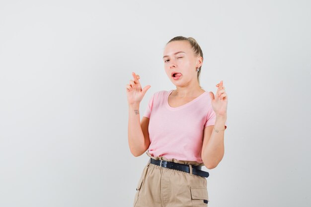 Chica rubia manteniendo los dedos cruzados, guiñando el ojo, sacando la lengua en la camiseta, vista frontal de los pantalones.