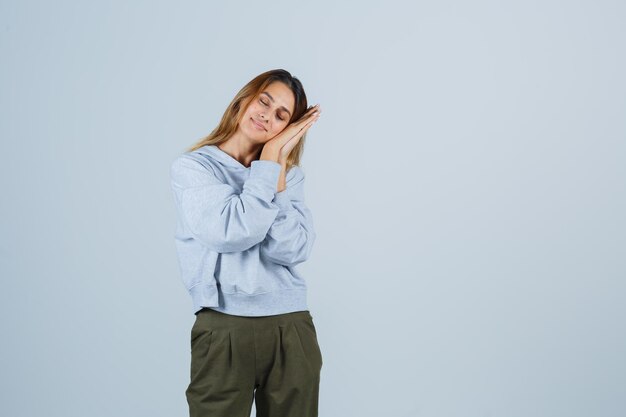 Chica rubia con la mejilla apoyada en las manos, fingiendo dormir en pantalones y sudadera azul verde oliva y con sueño. vista frontal.