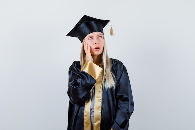 Chica rubia con la mejilla apoyada en la palma, mirando a otro lado en toga y gorra de graduación y mirando pensativo