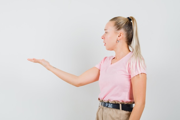 Chica rubia mirando atentamente su palma se extendió a un lado en camiseta, pantalones, vista frontal.