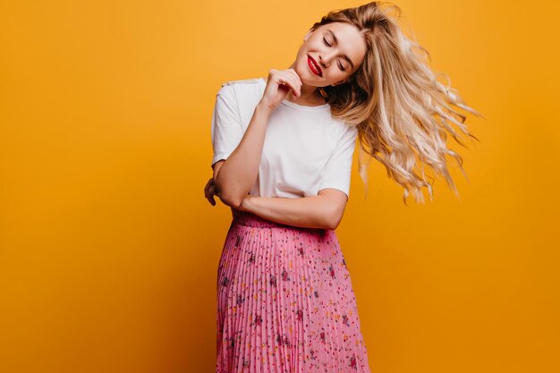 Chica rubia de moda disfrutando de la sesión de fotos Atractiva joven en camiseta blanca de pie sobre fondo amarillo