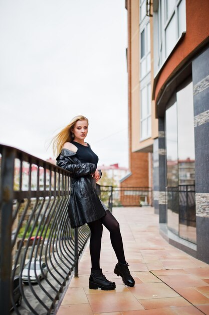 Chica rubia de moda con un largo abrigo de cuero negro posado contra una gran ventana del edificio