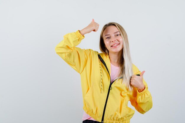 Chica rubia mostrando doble pulgar hacia arriba en camiseta, chaqueta y mirando alegre.