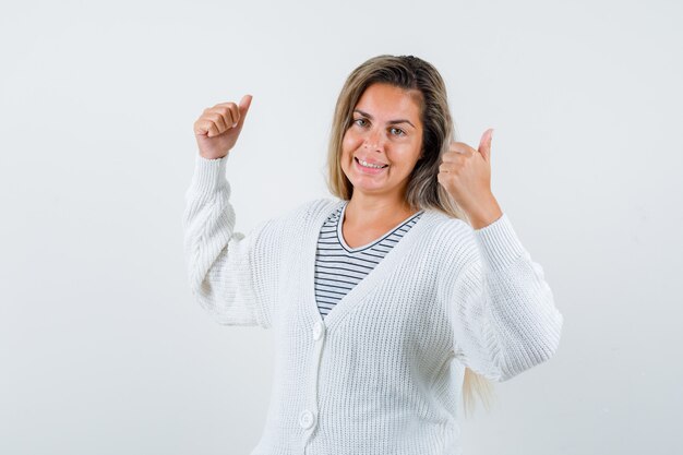 Chica rubia mostrando doble pulgar hacia arriba en camiseta a rayas, cárdigan blanco y pantalones de jean y mirando alegre, vista frontal.