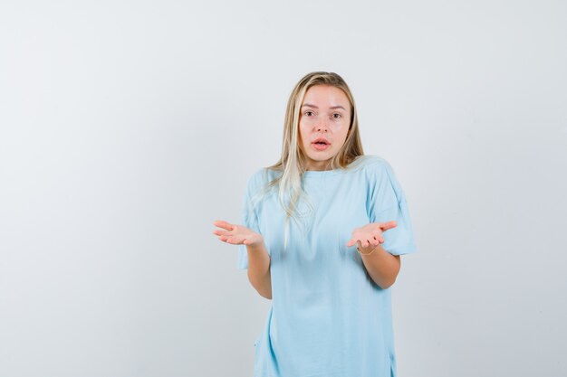 Chica rubia mostrando gesto de impotencia en camiseta azul y mirando confundido. vista frontal.