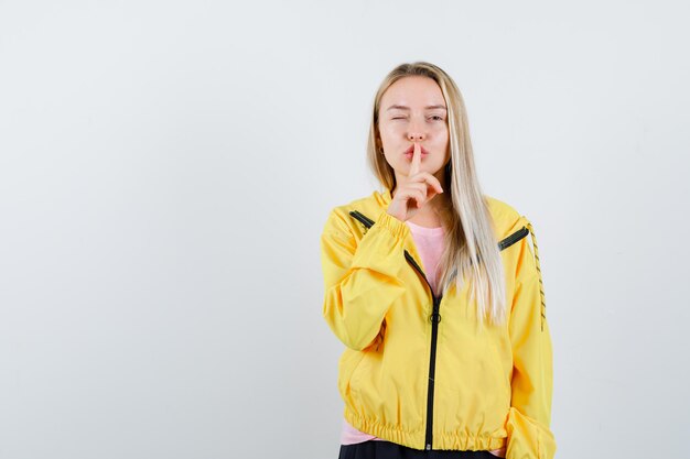 Chica rubia mostrando gesto de silencio en chaqueta amarilla y mirando confiado