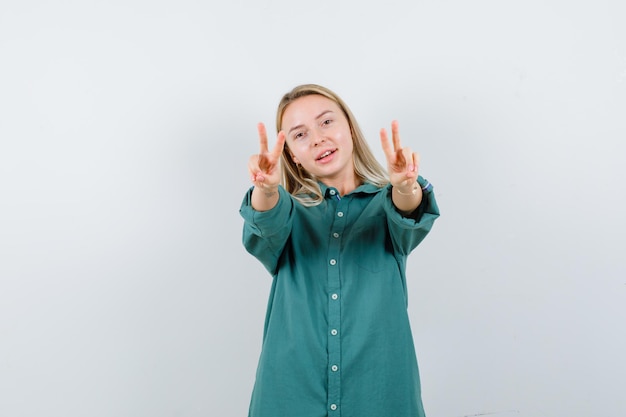 Chica rubia mostrando gestos de paz en blusa verde y mirando feliz