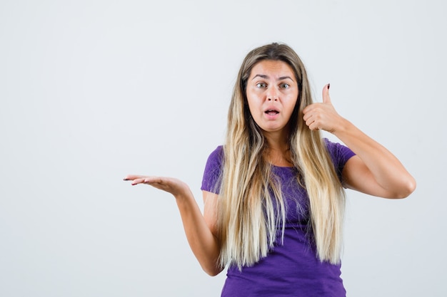 Chica rubia mostrando el pulgar hacia arriba mientras extiende la palma hacia un lado en la vista frontal de la camiseta violeta.