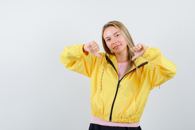 Chica rubia mostrando los pulgares hacia abajo con ambas manos en camiseta rosa y chaqueta amarilla y mirando feliz