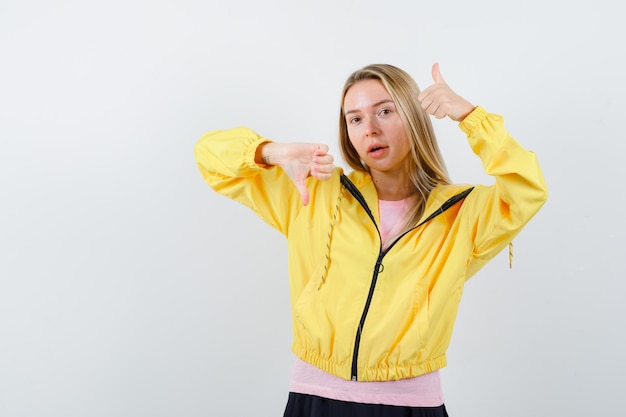 Chica rubia mostrando los pulgares hacia arriba y hacia abajo con ambas manos en camiseta rosa y chaqueta amarilla y mirando seria