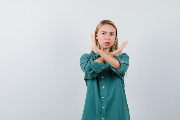 Chica rubia mostrando restricción o gesto x en blusa verde y mirando seria.