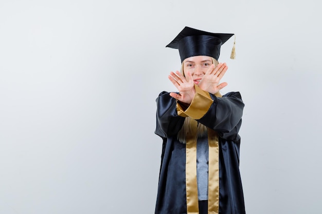 Foto gratuita chica rubia mostrando restricción o gesto x en toga y gorra de graduación y mirando seria