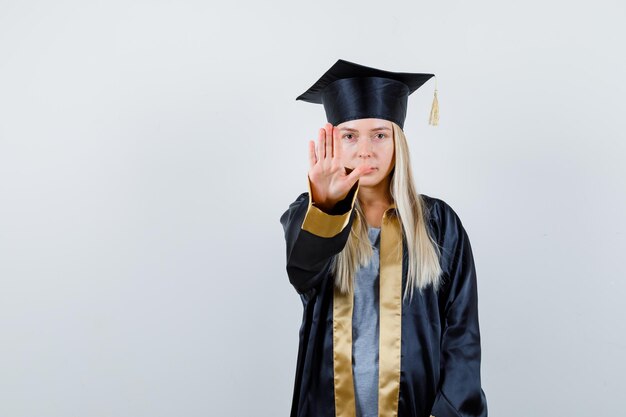 Chica rubia mostrando señal de stop en toga y gorra de graduación y mirando seria.