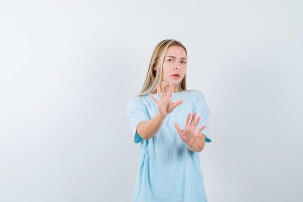 Chica rubia mostrando señales de alto en camiseta azul y mirando incómodo, vista frontal.