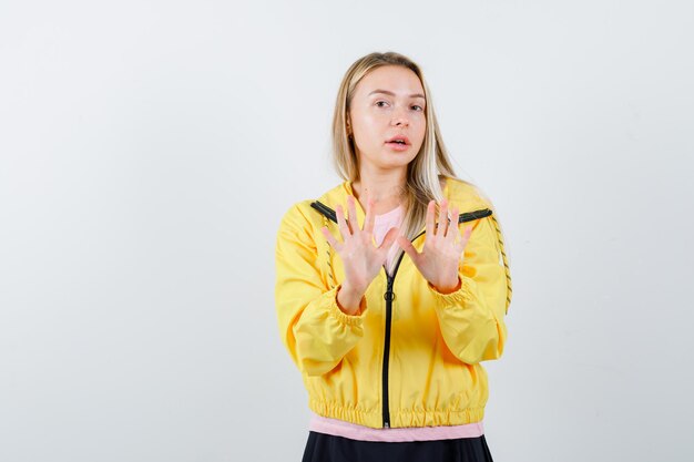 Chica rubia mostrando señales de alto en camiseta rosa y chaqueta amarilla y mirando asustada.