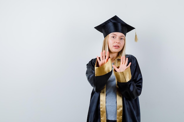 Chica rubia mostrando señales de alto en toga y gorra de graduación y mirando seria.