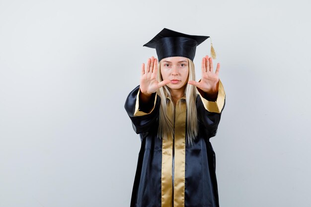 Chica rubia mostrando señales de alto en toga y gorra de graduación y mirando seria.