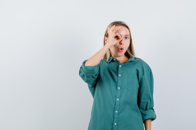 Chica rubia mostrando signo bien en el ojo en blusa verde y mirando sorprendido.